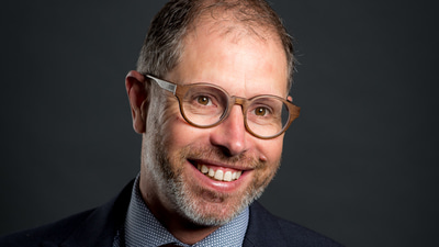 Nick has a bright smile and is wearing a navy suit and checked shirt with a spotted red tie and brown glasses