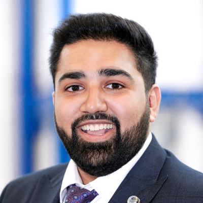 Hamza is photographed at an event. He smiles cheerfully at the camera. He wears a navy suit jacket, white shirt and patterned tie
