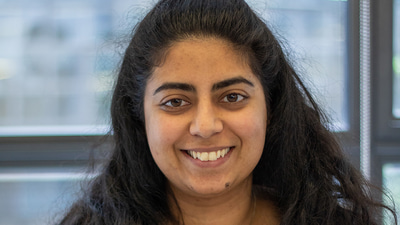 Deepali is seated in the AOP offices leaning her arms on a high table. She has long, dark curling hair, glasses, and a beige top