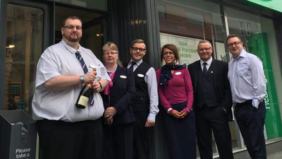 A group of six people stand outside of a Specsavers storefront, one holds a bottle of champagne