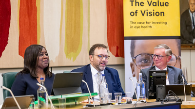 Three people sit at a long table, microphoens and screens in front of them. They are dressed in formal business attire. A banner in yellow and blacjk behind them states The value of vision, the case for investing in eye health