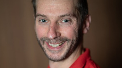Luke wears a red polo shirt and turns sideways to face the camera, smiling
