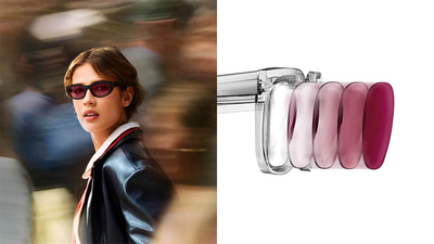 A woman pauses in a busy, blurred environment, to look at the camera. She wears black oval sunglasses with ruby lenses. To the right half is a representation of the Transition technology in the lens