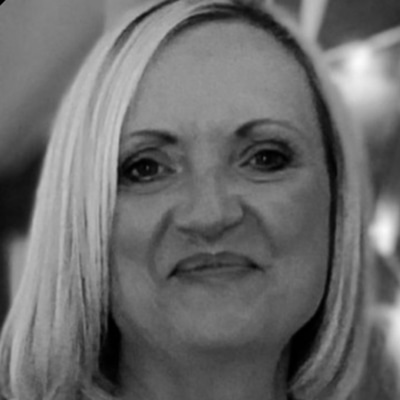 Black and white image shows a middle aged woman with shoulder length blonde hair smiling at the camera