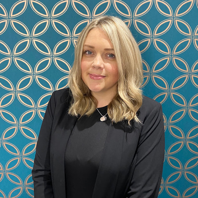A young woman dressed in a black suit and with shoulder length blonde hair is looking directly at the camera and smiling in front of royal blue wallpaper with a concentric gold pattern