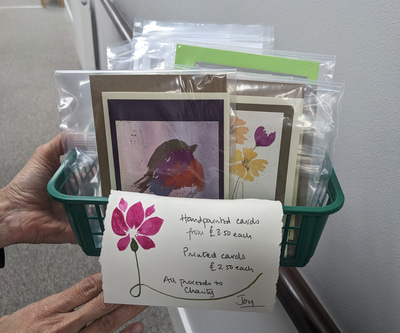 Two hands holding a basket of hand painted greeting cards. The first card visible is a painting of a robin. 