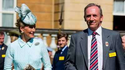 Nick Caplin smiling wearing a suit, standing next to a woman in a blue hat and suit