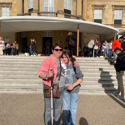 Charmaine and her daughter are posing with arms around each other in front of Buckingham Palace. Charmaine wears sunglasses, a pink jacket, and carries a white cane