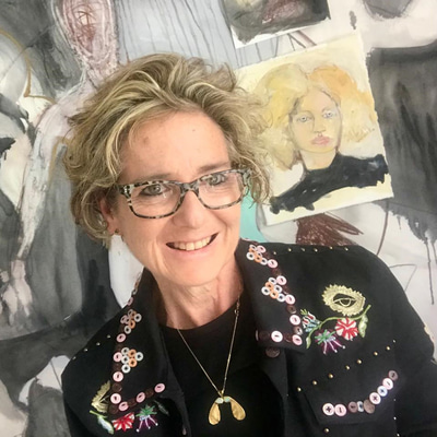 Rachel Gadsden wears a chunky rectangular frame with tortoiseshell pattern. She wears a dark jacket, embroidered with flowers and with pearlescent buttons sewn to the collar, and a necklace of ash seeds. Behind her can be seen a wall filled with layers of artwork.  