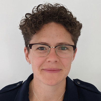Woman smiling with short brown curly hair. She is wearing a black shirt and glasses