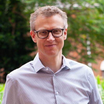 middle aged man smiling with dark grey hair. He is wearing an off-white shirt and glasses 