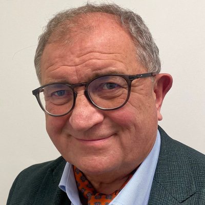 Sam smiles at the camera wearing grey-brown spectacles, a tweed suit and cheerful patterned orange tie