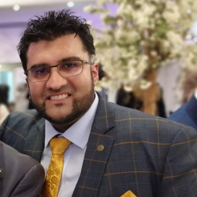 A man with black hair and a beard, smiling. He is wearing glasses and is dressed in a suit.