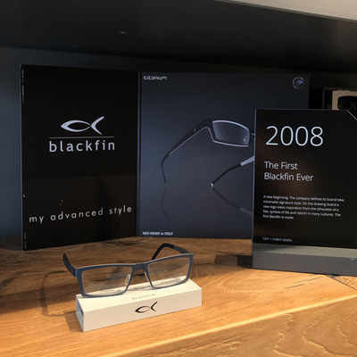 A black metal plaque reads: ‘2008. The first Blackfin ever.’ Behind it is a simple one page catalogue. The frame, a super-thin rectangular frame in grey metal is balanced on a little white prop with the Blackfin fish logo