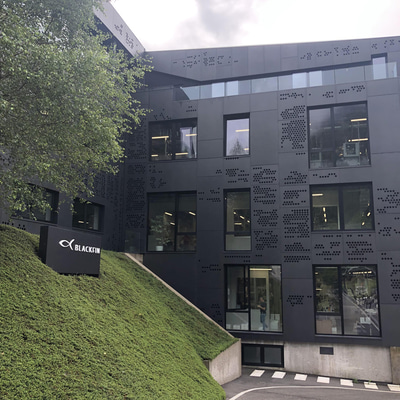 The black metal building has patterns of circular dots and many panes of long glass windows. On a steep green slope, a black sign has white font reading Blackfin and the fish logo