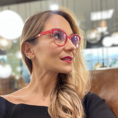 Carolina is seated in a restaurant, looking off to the side, with bright red cat’s-eye style glasses 