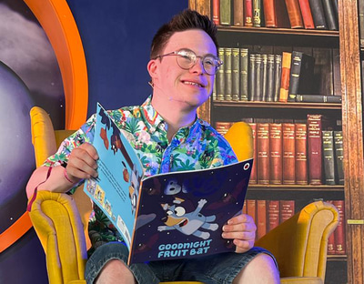 George wears circle spectacles with his hair gelled up and a colourful Hawaiian shirt. He is sitting in a bright yellow armchair and holding a Bluey book called ‘Goodnight fruit bat.’