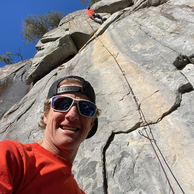 Wearing white sunglasses with purple polarised lenses and a backwards cap, Tim takes a selfie from the bottom of a rockface. Another climber is high above.