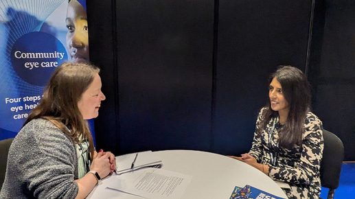 Emily, left, and Priya, right, are seated around a circle table for an interview. On the table are notes, notebooks, copies of OT’s guides and in the background is a banner for the AOP
