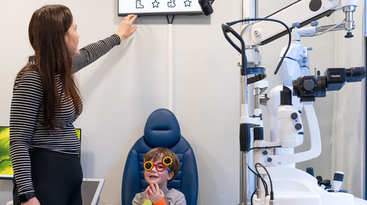 Fiona points to an eye test chart that uses icons of a star, duck, apple, boot, instead of letters. An infant boy in the consulting chair wears an occluder with one lens over his eye and is looking ahead