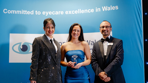Three people pose in front of a blue backdrop that reads “Committed to eyecare excellence in Wales.” The woman in the centre, wearing a blue strapless gown, holds a small glass award and smiles. To her left stands a woman in a sparkly black suit holding a microphone, and to her right stands a man in a black tuxedo. All three are dressed formally and smiling under bright event lighting