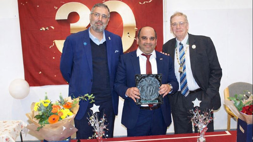 Three men in smart suits stand behind a red table covered with flowers and decorations. The man in the centre holds a glass award. Behind them is a red banner with a gold 30
