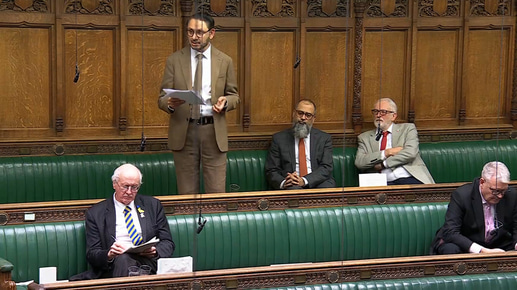 A wide view of the House of Commons chamber shows a man standing on the green benches reading from papers as he speaks, with several other MPs seated nearby listening or reading, against the backdrop of carved wooden panelling and hanging microphones