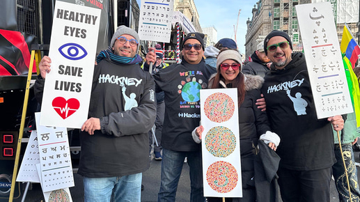 Three men and one woman cheer and raise placards in the New Years Day parade. The placards include sight tests in different language scripts, a sign for ‘Healthy eyes save lives’ and a three colour vision tests. The people are wrapped up warmly, and over their coats they wear t-shirts celebrating Hackney and unity