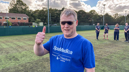 A man in a blue SpaMedica branded t-shirt and cataract simulation goggles holds a thumb up
