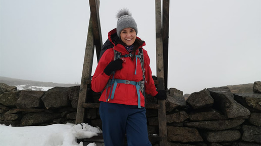 A woman wearing a red jacket, blue trousers, gloves, and a grey bobble hat stands on a snow-covered wooden stile built into a stone wall. She smiles at the camera, surrounded by deep snow and a foggy, white sky