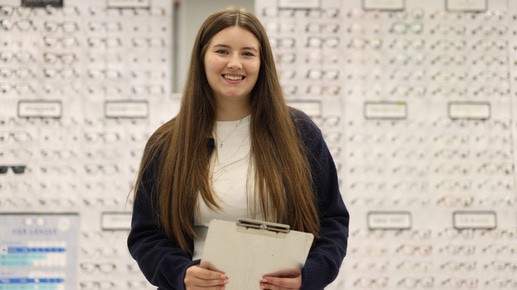 A woman stands smiling in front of a large display wall filled with spectacle frames. She is holding a clipboard and wearing a white top with a dark cardigan 