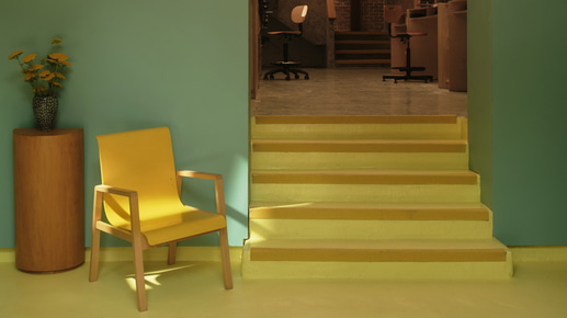 A small reception space with a soft yellow floor, yellow stairs, and a half-wall painted in teal. There is a wooden yellow chair, with shelving and flowers beside it. The stairs lead to a workshop, built in wood against brick walls, while postmodern stairs in grey concrete rise up behind under an arching glass ceiling