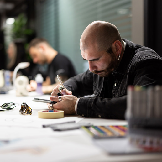 A designer, wearing a dark denim jacket and many rings, is sketching on a tablet. He is surrounded by papers of eyewear designs and has several frames around him