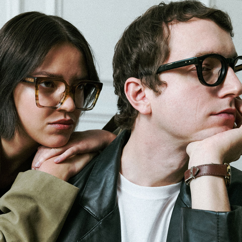 From a photoshoot – a woman leans against a man’s shoulder, with her chin resting on her hands. She wears oversized square acetate frames in shades of brown. The man, in a black leather jacket, wears black frames. 