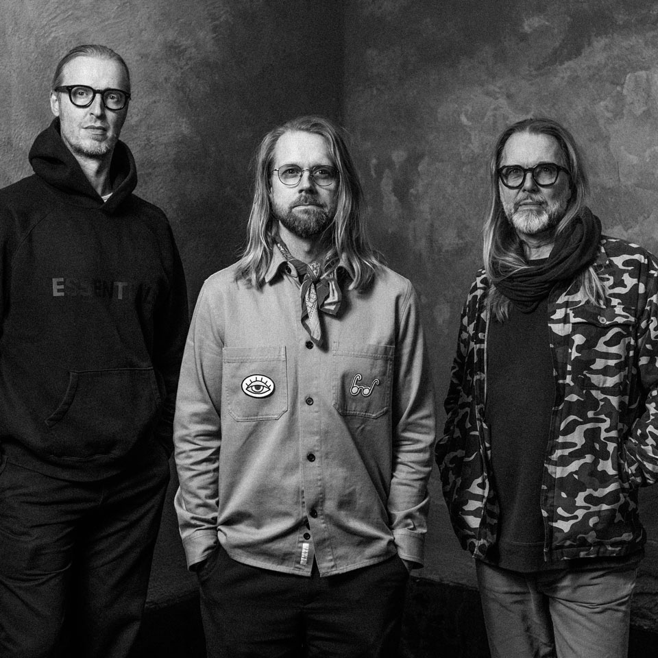 A black and white image of three men standing in a line with their hands in their pockets. Their expressions are curious but unsmiling. They all have long hair, one tied back. Two wear thick frames and a third wears thin metal spectacles