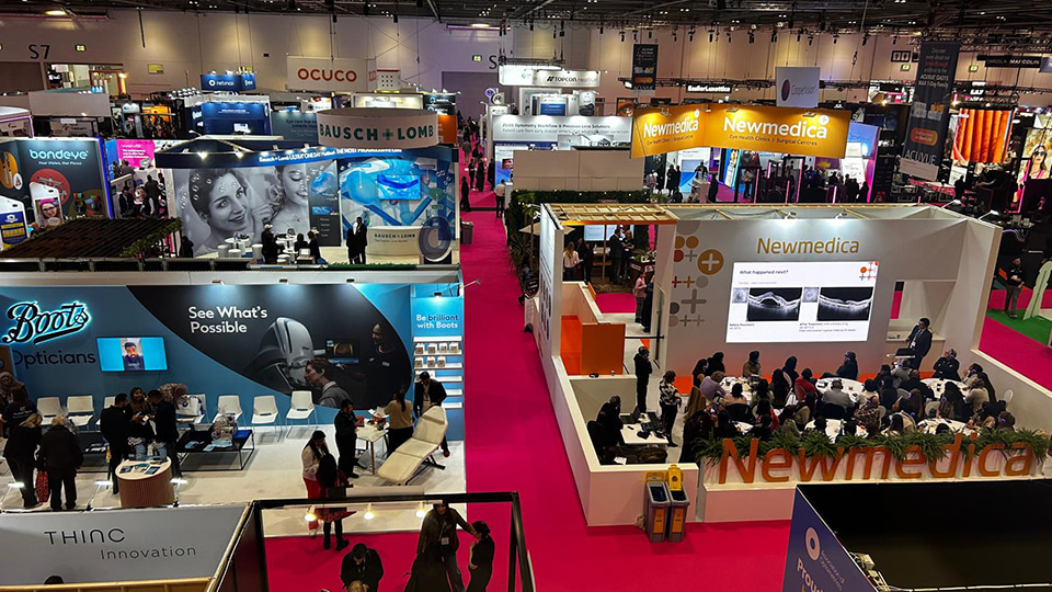 High-angle view of a busy optical industry exhibition hall with bright pink carpeted aisles and large branded stands, including Boots Opticians, Bausch + Lomb, and Newmedica. Attendees walk between booths, sit in meeting areas, and watch presentations on large screens while company displays showcase eye care technology and services