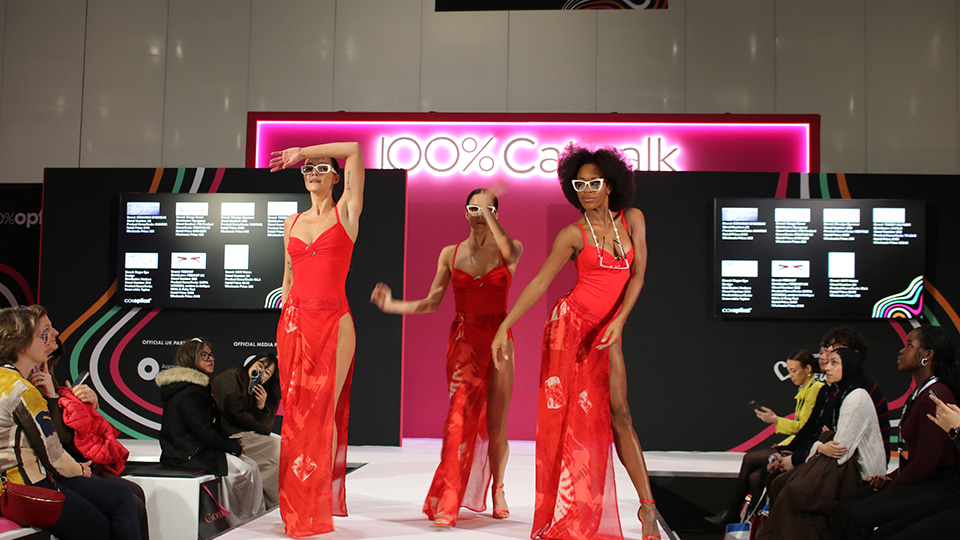 Three models wearing white sunglasses and bright red dresses pose and dance on a runway at the “100% Catwalk” stage, with audience members seated on both sides watching the eyewear fashion show.