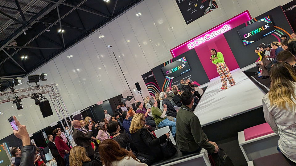 A presenter in a bright green top and patterned trousers speaks on the “100% Catwalk” stage at an optical trade show, while a large seated audience watches from benches around the runway