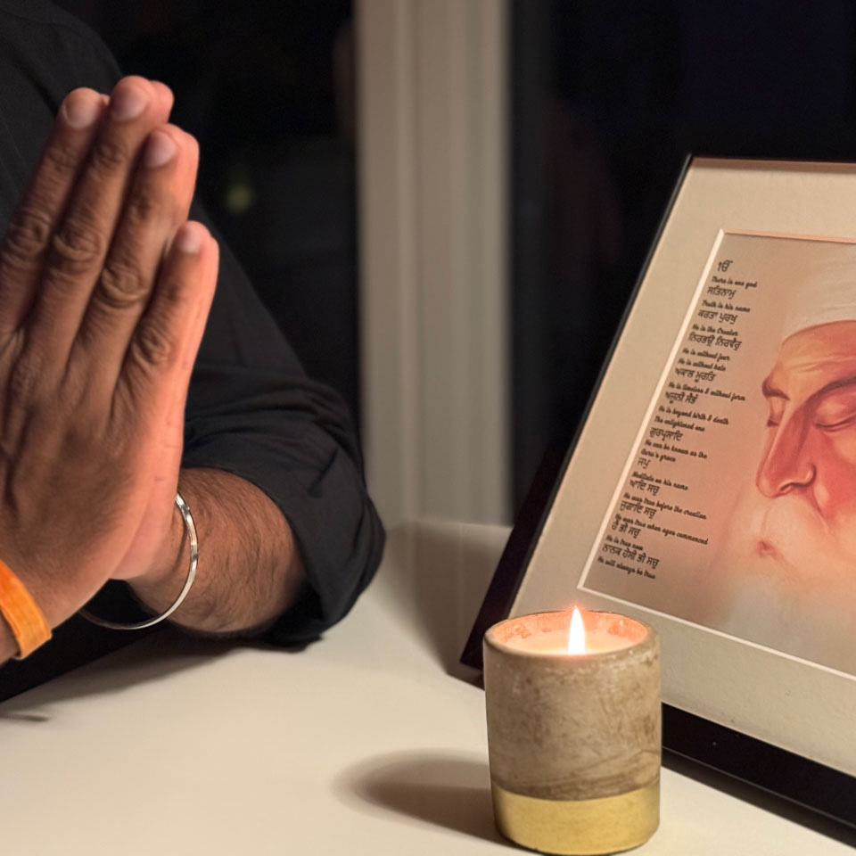 Two hands pressed together by a candle, the wrist of one is wrapped in an orange band. A metal bracelet circles the other. 