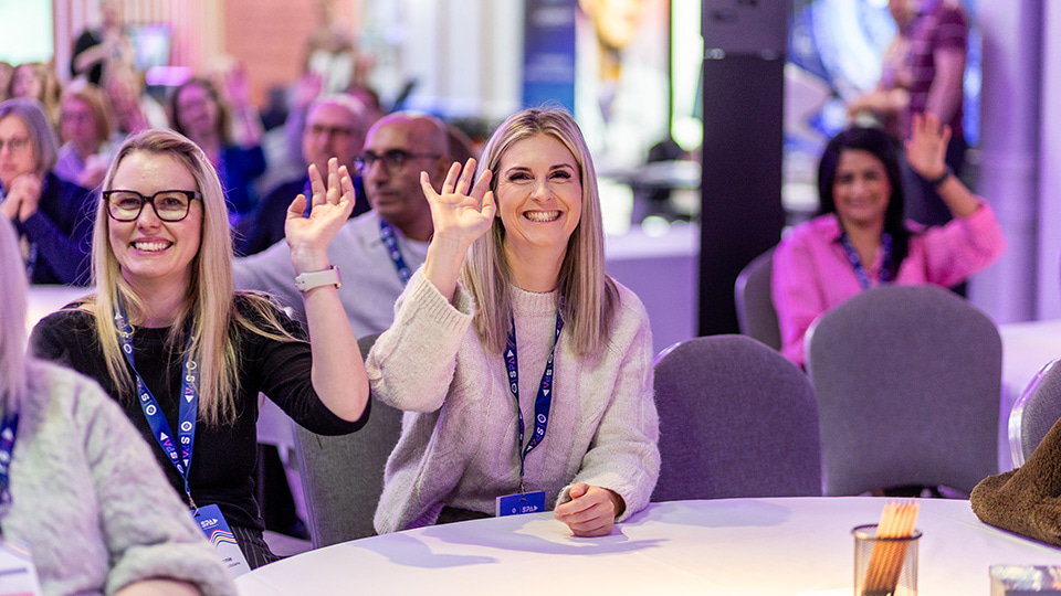 Attendees seated at round tables during a conference session smile and raise their hands to answer a question. Two women in the foreground wearing event lanyards wave enthusiastically, while other delegates participate in the background under purple and blue stage lighting