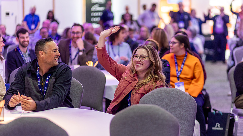 A conference audience engages in an interactive session, with a woman in the foreground raising her hand while smiling. Fellow attendees seated around round tables, wearing event lanyards, watch the presentation and chat as the room is lit with soft purple and warm ambient lighting