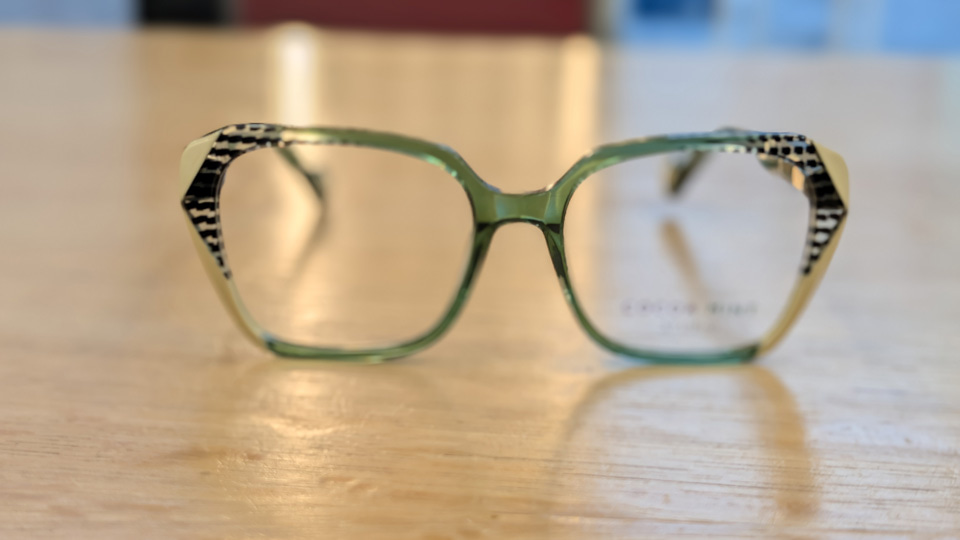 An oversized but thin pair of spectacles with clear green on three sides of the frame around the brown, bridge, and bottom. On either edge, the frame has a slight cat’s eye lift, and changes to a creamy opqaue light breen, intercut with black and white patterns