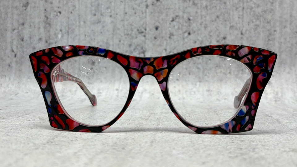 A pair of large spectacles in a geometric shape similar to a cat’s eye but much more squared. The frame itself is made up of irregular small pieces of acetate in reds and purples, joined together by black in an effect similar to stained glass