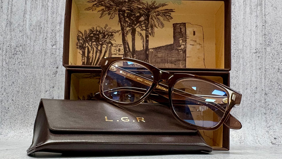 A pair of brown square spectacles rest on the top of a soft leather case with L.G.R in gold lettering. Behind them both is the open box, with an illustration of an old building and palm trees. The effect is classic and classy