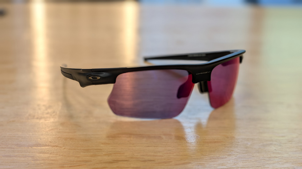 A pair of sports sunglasses in black with rose coloured lenses. The arms are attached directly to the browbar creating one curving line and leaving the bottom of the lenses as rimless
