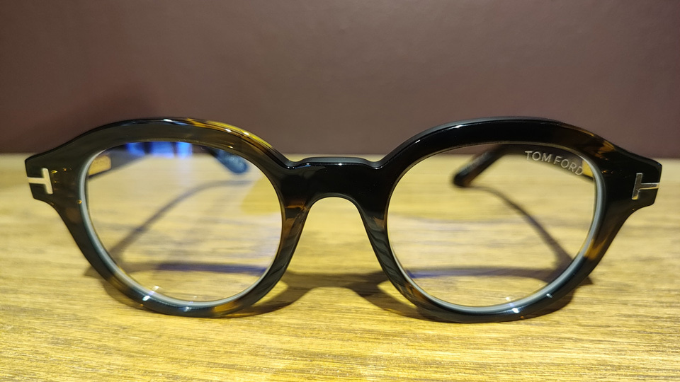 A pair of chunky rounded spectacles in a really dark acetate, with lighter and darker areas, A metal ‘T’ on the sides indicate this is Tom Ford. The effect is bold but not overpowering