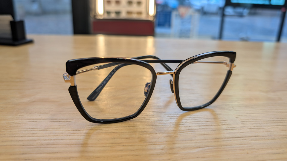 A thin cat’s eye style pair of spectacles with slimline black frame front. The black acetate of the frame front stops either side of the hinge, shaped as a silver ‘T’ representing the brand, which gives an added interest to the look. The metal rims around the frame front are clearly visible
