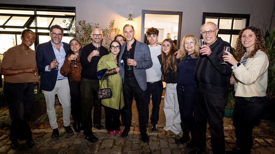 A group of 12 people gather outside of a building. They all grin and raise champagne glasses in a toast. Everyone is dressed smartly