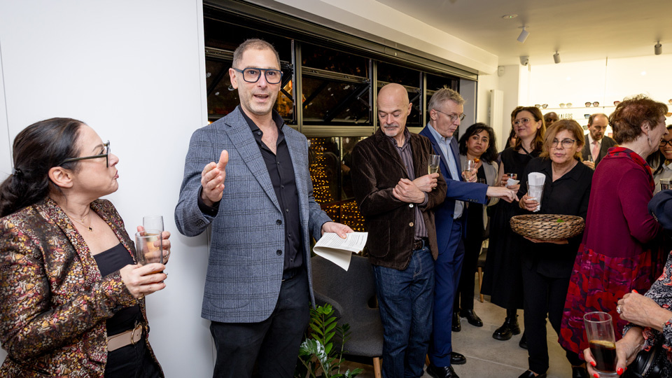 Jonathan Cohen in black shirt and dark jeans with grey tweed jacket and black aviator spectacles gives a speech to a gathered crowd, all wearing party clothes
