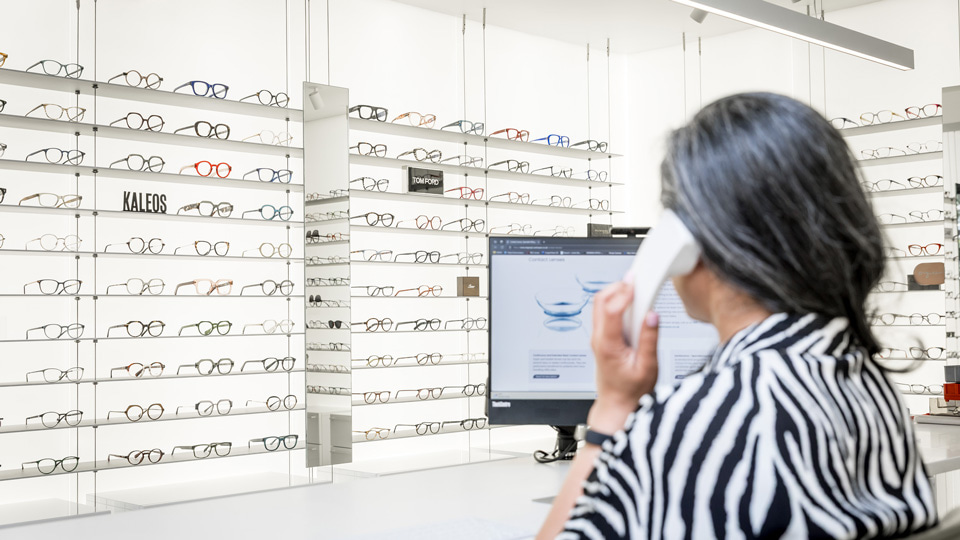 The image is taken from behind a staff member who is on the phone, contact lenses displayed on her computer screen. Across the room from her are two bright white walls filled with simple, minimalist shelves of bright, colourful frames and mirrors